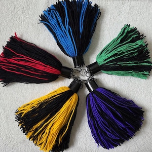 Op de afbeelding: Vijf kleurrijke garen kwasten in een cirkelvormig patroon. Elke kwast heeft een zwarte basis met een contrasterende kleur: rood, blauw, groen, geel en paars. De kwasten zijn bevestigd aan zwarte metalen doppen met sleutelringen.