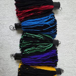 Op de afbeelding: Vijf kleurrijke kwasten met zwart, rood, blauw, groen, paars en geel garen. Elke kwast heeft een zwarte cilindrische bovenkant met een metalen lus. De kwasten zijn verticaal gerangschikt tegen een witte, getextureerde achtergrond.