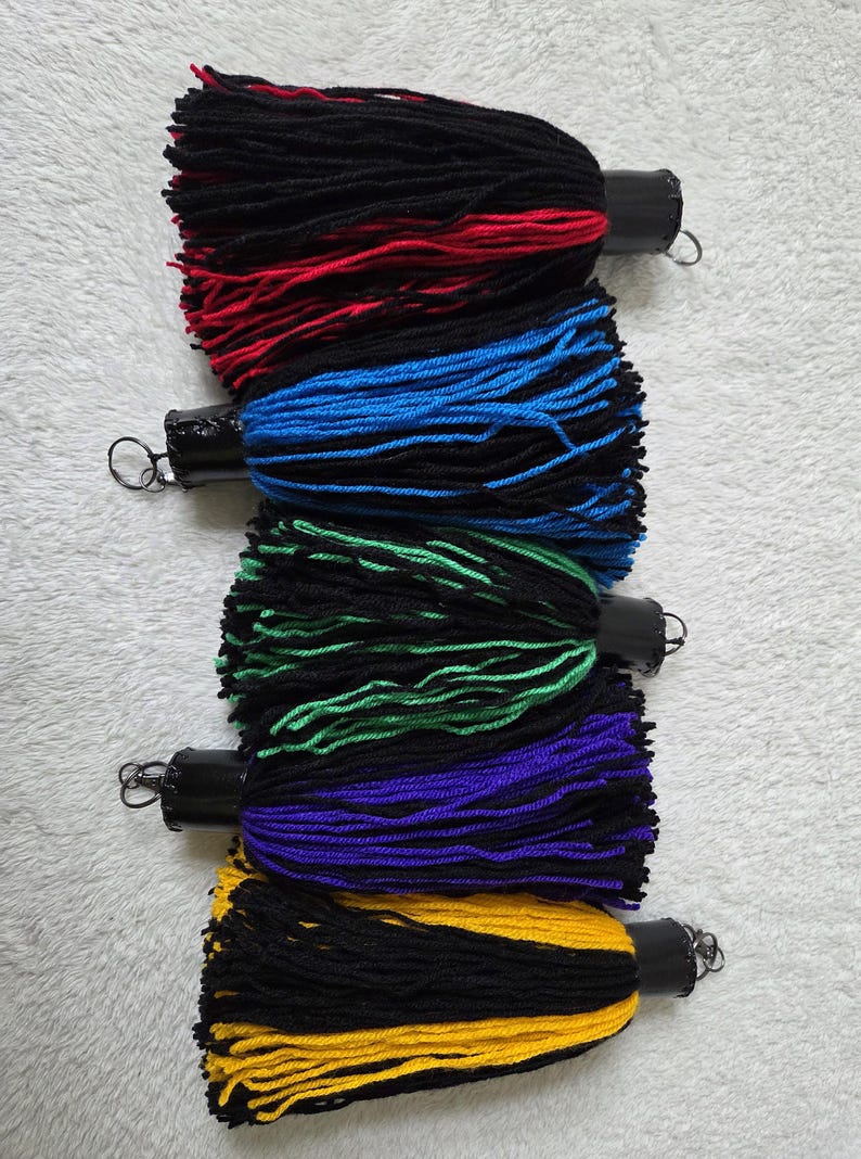 Op de afbeelding: Vijf kleurrijke kwasten met zwart, rood, blauw, groen, paars en geel garen. Elke kwast heeft een zwarte cilindrische bovenkant met een metalen lus. De kwasten zijn verticaal gerangschikt tegen een witte, getextureerde achtergrond.