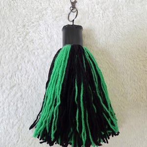 Op de afbeelding: Een handgemaakte kwast sleutelhanger met een zwarte cilindrische bovenkant en een mix van groene en zwarte garenstrengen. De sleutelhanger bevat een zilverkleurige sluiting voor bevestiging.