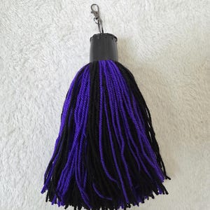 Op de afbeelding: Een handgemaakte kwastjes sleutelhanger met een zwarte bovenkant en een mix van zwarte en paarse garenstrengen. De sleutelhanger heeft een zilverkleurige sluiting voor eenvoudige bevestiging aan sleutels of tassen. De kwast is een decoratief accessoire.