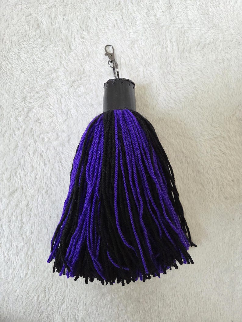 Op de afbeelding: Een handgemaakte kwastjes sleutelhanger met een zwarte bovenkant en een mix van zwarte en paarse garenstrengen. De sleutelhanger heeft een zilverkleurige sluiting voor eenvoudige bevestiging aan sleutels of tassen. De kwast is een decoratief accessoire.