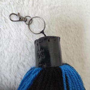 Op de afbeelding: Een handgemaakte sleutelhanger met een zwarte en blauwe kwast. De kwast is bevestigd aan een zwart met leer omwikkeld bovenstuk met een zilverkleurige sluiting en sleutelring. De kwast is gemaakt van blauw en zwart garen.