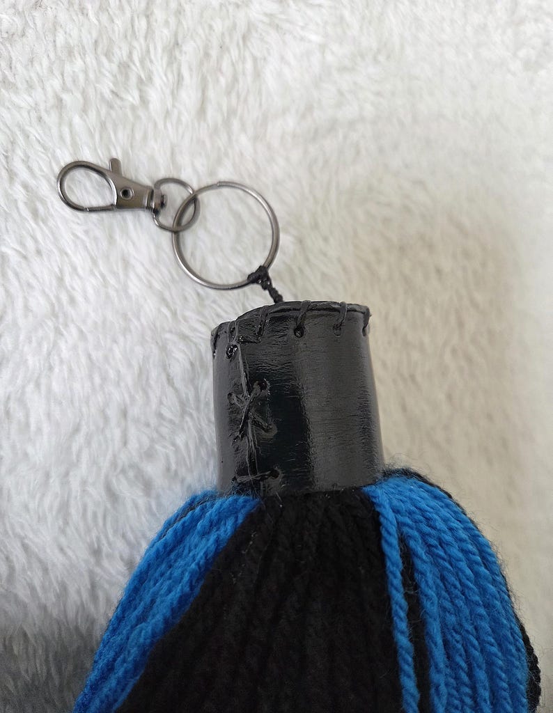 Op de afbeelding: Een handgemaakte sleutelhanger met een zwarte en blauwe kwast. De kwast is bevestigd aan een zwart met leer omwikkeld bovenstuk met een zilverkleurige sluiting en sleutelring. De kwast is gemaakt van blauw en zwart garen.