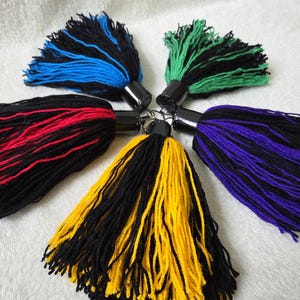 Op de afbeelding: Een verzameling van zes kleurrijke kwasten, elk met een zwarte cilindrische bovenkant. De kwasten hebben combinaties van zwart met blauw, groen, rood, geel en paars. De kwasten zijn gemaakt van garen en zijn in een cirkelvorm gerangschikt.