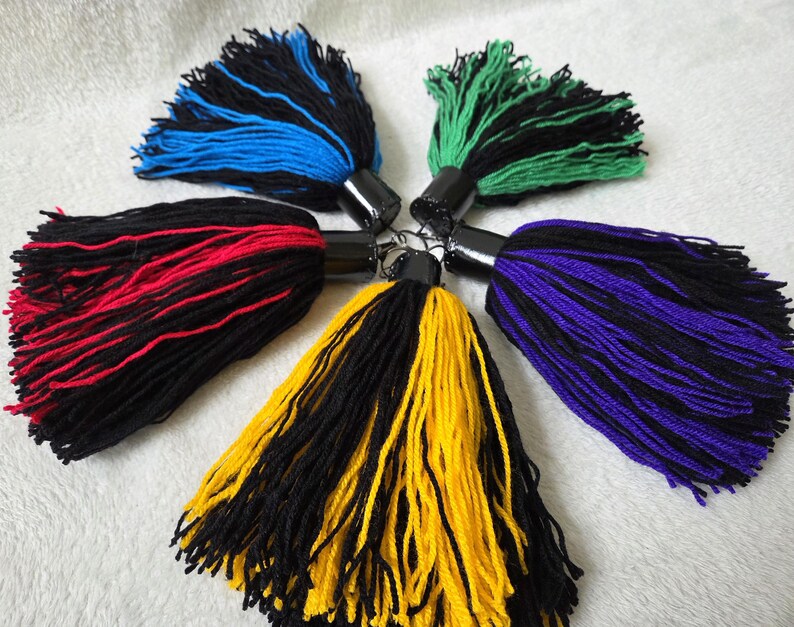 Op de afbeelding: Een verzameling van zes kleurrijke kwasten, elk met een zwarte cilindrische bovenkant. De kwasten hebben combinaties van zwart met blauw, groen, rood, geel en paars. De kwasten zijn gemaakt van garen en zijn in een cirkelvorm gerangschikt.