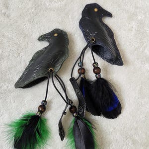 May include: Two raven-shaped leather ornaments. Each raven is black with a gold-colored eye and a leather strap. The ornaments have beaded accents and are adorned with black, green, and blue feathers. The background is a white, textured surface.