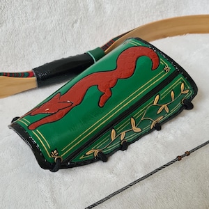 May include: Green and black leather archery arm guard with a red fox design. The arm guard has gold accents and a decorative leaf pattern. A bow and arrow are also visible.