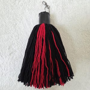 Op de afbeelding: Een handgemaakte sleutelhanger met kwast, met een zwart en rood garen ontwerp. De kwast is bevestigd aan een zwarte cilindrische dop met een zilverkleurige sluiting. De garenstrengen zijn lang en hangen naar beneden.