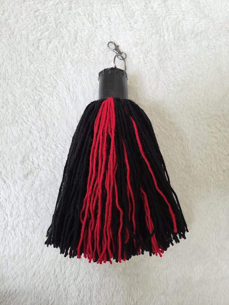 Op de afbeelding: Een handgemaakte sleutelhanger met kwast, met een zwart en rood garen ontwerp. De kwast is bevestigd aan een zwarte cilindrische dop met een zilverkleurige sluiting. De garenstrengen zijn lang en hangen naar beneden.