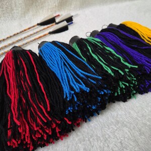 Op de afbeelding: Een verzameling kleurrijke garen kwasten in rood, blauw, groen, paars en geel, met zwarte bases. Drie pijlen met witte en zwarte veren zijn zichtbaar op de achtergrond. De kwasten zijn gerangschikt op een witte ondergrond.