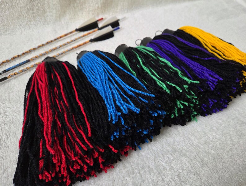 Op de afbeelding: Een verzameling kleurrijke garen kwasten in rood, blauw, groen, paars en geel, met zwarte bases. Drie pijlen met witte en zwarte veren zijn zichtbaar op de achtergrond. De kwasten zijn gerangschikt op een witte ondergrond.