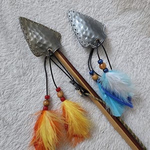 May include: Two decorative arrows with silver-colored arrowhead tips and wooden shafts. Each arrow features colorful feather accents in orange, yellow, and blue, along with wooden and blue bead details. The arrows are designed for display.