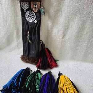 Op de afbeelding: Zwarte koker met bruine bontafwerking en decoratieve patches van een wolf, een beer en een kompasachtig symbool. De koker heeft een rits en is versierd met kwasten in verschillende kleuren, waaronder blauw, groen, paars en geel.
