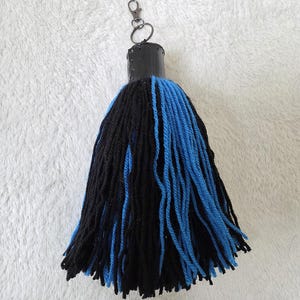 Op de afbeelding: Een handgemaakte sleutelhanger met een zwarte en blauwe kwast. Het heeft een zwarte cilindrische bovenkant met een metalen sluiting en ring. De kwast is gemaakt van dik garen, met afwisselende secties van zwarte en blauwe strengen.