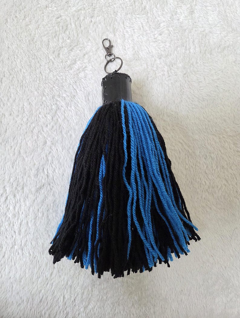 Op de afbeelding: Een handgemaakte sleutelhanger met een zwarte en blauwe kwast. Het heeft een zwarte cilindrische bovenkant met een metalen sluiting en ring. De kwast is gemaakt van dik garen, met afwisselende secties van zwarte en blauwe strengen.
