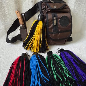 Op de afbeelding: Een bruine crossbodytas met een zwarte riem, een mes in een foedraal en verschillende kleurrijke kwasten. De kwasten zijn zwart en geel, rood, blauw, groen en paars.
