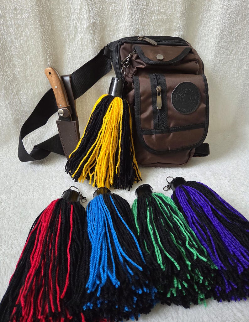 Op de afbeelding: Een bruine crossbodytas met een zwarte riem, een mes in een foedraal en verschillende kleurrijke kwasten. De kwasten zijn zwart en geel, rood, blauw, groen en paars.