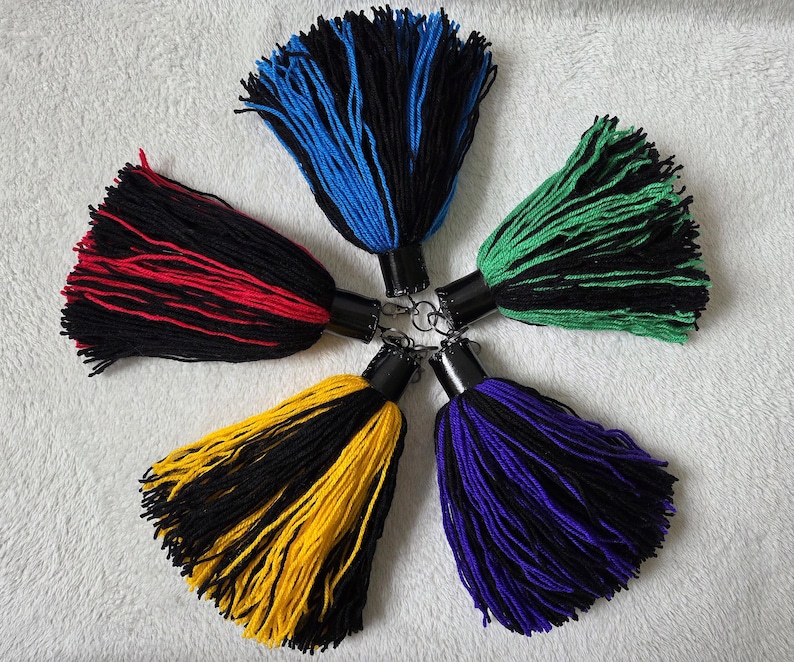 Op de afbeelding: Vijf kleurrijke garen kwasten in een cirkelvormig patroon. Elke kwast heeft een zwarte basis met een contrasterende kleur: rood, blauw, groen, geel en paars. De kwasten zijn bevestigd aan zwarte metalen doppen met sleutelringen.