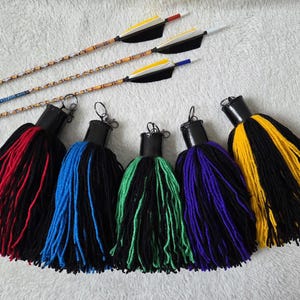 Op de afbeelding: Een assortiment boogschietaccessoires. Vijf grote kwasten in rood, blauw, groen, paars en geel met zwarte accenten worden getoond onder drie pijlen met decoratieve schachten en veren. De pijlen hebben zwarte schachten en gekleurde veren.