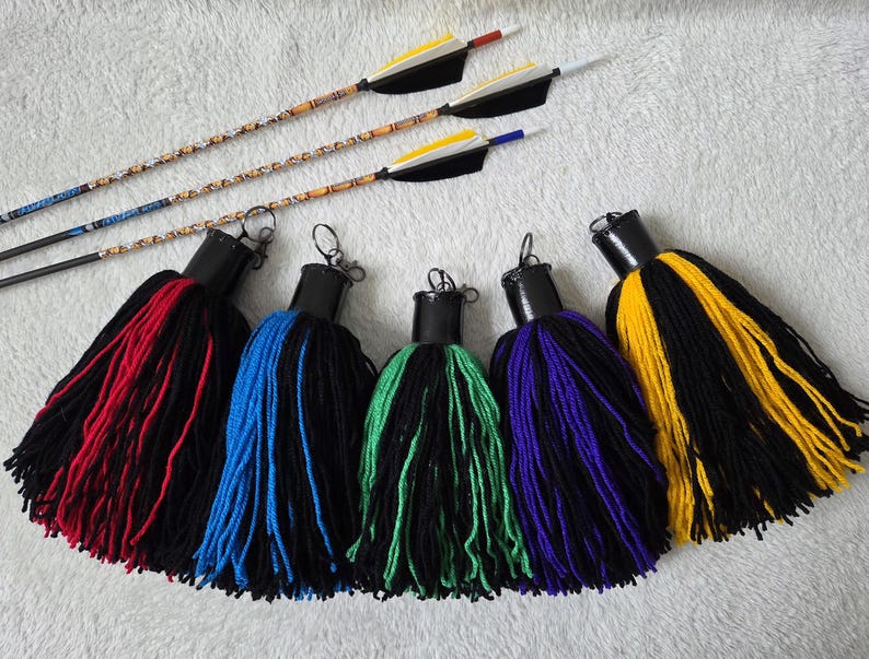 Op de afbeelding: Een assortiment boogschietaccessoires. Vijf grote kwasten in rood, blauw, groen, paars en geel met zwarte accenten worden getoond onder drie pijlen met decoratieve schachten en veren. De pijlen hebben zwarte schachten en gekleurde veren.