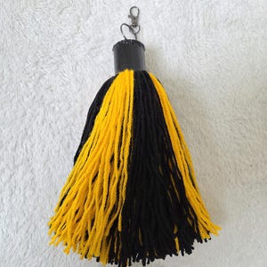 Op de afbeelding: Een handgemaakte kwast-sleutelhanger met een zwarte bovenkant en een mix van gele en zwarte garenstrengen. De sleutelhanger heeft een metalen sluiting voor eenvoudige bevestiging aan sleutels of tassen. Een decoratief accessoire.