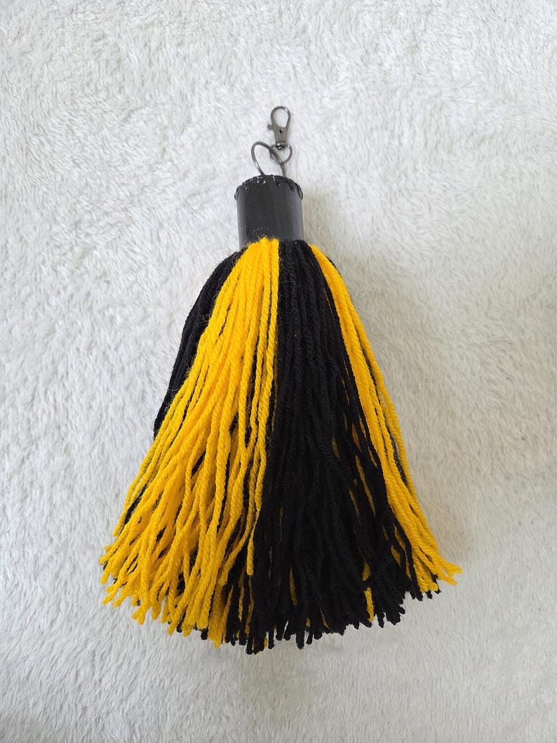 Op de afbeelding: Een handgemaakte kwast-sleutelhanger met een zwarte bovenkant en een mix van gele en zwarte garenstrengen. De sleutelhanger heeft een metalen sluiting voor eenvoudige bevestiging aan sleutels of tassen. Een decoratief accessoire.