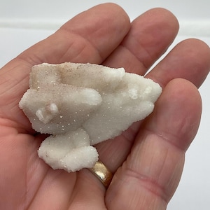 May include: A cluster of white and tan crystals, possibly quartz or a similar mineral, held in a hand. The crystals have a textured, granular appearance and are illuminated by soft lighting. The overall shape is irregular and organic.