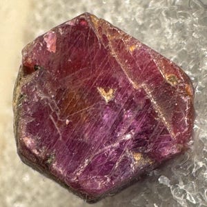 May include: A faceted, raw ruby gemstone with a deep red hue. The surface has natural lines and imperfections, with hints of lighter red and brown. The gemstone is set against a textured, light-colored background.