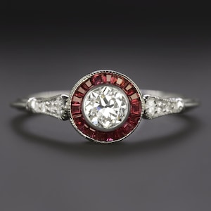May include: A vintage-style ring with a central, round, clear diamond surrounded by a halo of small, rectangular red gemstones. The band is silver-toned, with small accent diamonds on each side of the central stone. The ring is set against a dark background.