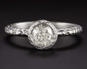 Könnte beinhalten: Ein silberner Ring mit einer strukturierten Band und einem runden Diamanten in einer Zargenfassung.