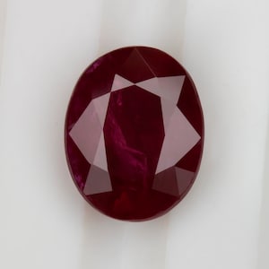 May include: A deep red ruby gemstone with a faceted cut. The gemstone is oval shaped and has a high polish.