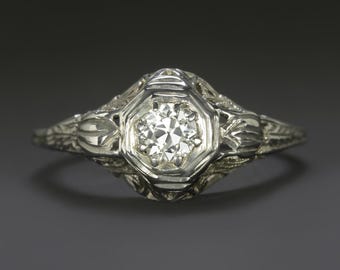 May include: A vintage-style engagement ring with a central, round-cut diamond set in an octagonal, white metal bezel. The band features intricate filigree detailing, with floral and leaf motifs. The ring is displayed against a dark gray background.