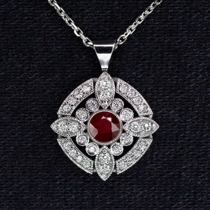 May include: A silver pendant necklace with a central, round, deep red gemstone, encircled by a detailed design of smaller, clear gemstones. The pendant hangs from a delicate silver chain, set against a dark background.