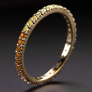 Può includere: Un anello d'oro con una fascia completa di pietre preziose. Le pietre preziose cambiano colore dal giallo all'arancione. L'anello è posto su uno sfondo scuro.