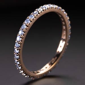 May include: Rose gold eternity ring featuring a continuous line of light blue gemstones. The ring is set with small, round-cut stones, creating a sparkling effect. The band is a warm, metallic colour, and the ring is displayed against a dark background.