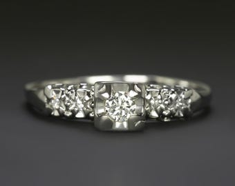 Può includere: Anello d'argento con pietra centrale quadrata e pietre più piccole ai lati. Lo sfondo grigio scuro esalta la brillantezza dei diamanti. Un design classico e senza tempo.