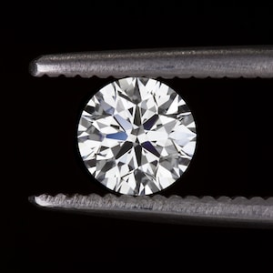 Pode incluir: Um diamante redondo lapidado em brilhante, segurado por uma pinça prateada contra um fundo preto. O diamante brilha com inúmeras facetas, refletindo a luz. A pinça está em foco, destacando a clareza da gema.