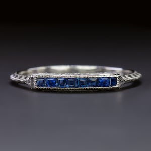 Könnte beinhalten: Ein zarter Platin-Bandring mit einer Reihe von sieben quadratischen blauen Saphiren, die in einer Kanalfassung gefasst sind. Das Band ist mit einem floralen Muster graviert.