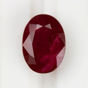 Pode incluir: Uma gema de rubi de corte oval com um tom vermelho profundo. A gema tem múltiplas facetas que refletem a luz, criando um efeito brilhante. O rubi está sobre um fundo branco.