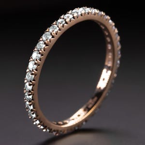 May include: A rose gold eternity ring featuring a continuous line of small, round, clear gemstones. The ring's band has a warm, metallic hue, and the gemstones are closely set, producing a sparkling effect. The ring is shown against a dark backdrop.