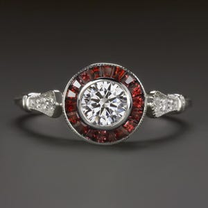 Puede incluir: Un anillo de plata con una piedra central redonda y transparente rodeada por un halo de piedras preciosas rojas. La banda está decorada con piedras preciosas más pequeñas y transparentes.