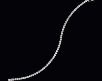 May include: A delicate, sparkling tennis bracelet against a black background. The bracelet features a continuous line of round-cut, clear gemstones, likely diamonds, set closely together in a silver or white gold setting. The clasp is visible at the end.