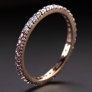 May include: Rose gold eternity ring with a gradient of gemstones. The ring features a row of small, round gemstones that transition from purple to light blue. The ring is set against a dark background.