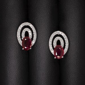 May include: A pair of oval-shaped ruby and diamond earrings. Each earring features a large, oval-cut ruby at the center, surrounded by a halo of small, sparkling diamonds. The diamonds are set in a silver or white gold setting.