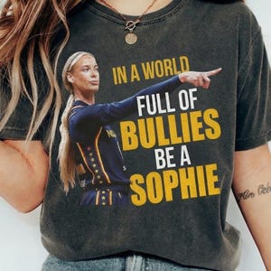 May include: A charcoal gray t-shirt featuring a graphic of a basketball player in a navy and gold jersey pointing to the right. The text reads IN A WORLD FULL OF BULLIES BE A SOPHIE in yellow and white lettering. The shirt has a crew neckline.