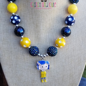 May include: A yellow and blue beaded necklace with a charm of a girl wearing a yellow raincoat. The necklace is on a mannequin and the text "Classy and Cute" is visible.