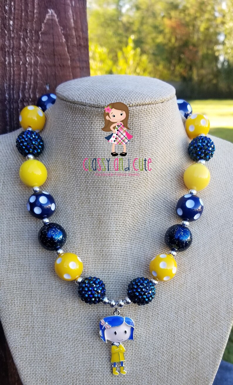 May include: A yellow and blue beaded necklace with a charm of a girl wearing a yellow raincoat. The necklace is on a mannequin and the text "Classy and Cute" is visible.