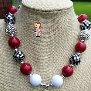 May include: A chunky necklace with red, white, silver, and black beads. The necklace features a pendant with the text "ALABAMA CRIMSON TIDE" around a large letter "A". The beads are displayed on a neutral-colored surface.