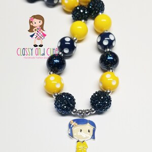 May include: A yellow and blue beaded necklace with a charm of a girl in a yellow raincoat. The necklace has white polka dots on the yellow beads and blue beads with glitter.